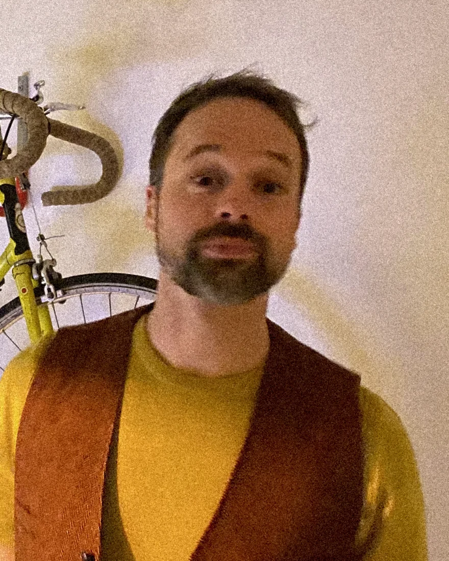 Portret van Remko Adema in geel shirt en roestbruin gilet, met een gele racefiets aan de muur